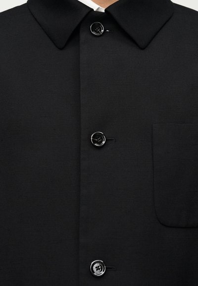 Μαύρο σακάκι με κουμπιά και μυτερό γιακά, που διαθέτει δύο μαύρα κουμπιά και επίπεδη τσέπη στην αριστερή πλευρά. Λείος υφασματικός, μινιμαλιστικός σχεδιασμός.