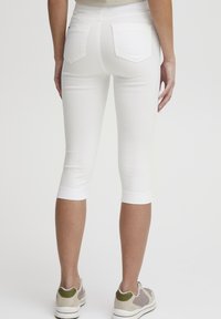 Leggings capri blancos con un diseño ajustado, que cuentan con bolsillos traseros y una textura suave, combinados con zapatillas deportivas de color claro.