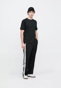 Čierne tričko s krátkym rukávom s textúrovanými detailmi, v kombinácii s čiernymi nohavicami s bielymi bočnými pruhmi a nášivkou s logom, doplnené čiernou čiapkou.