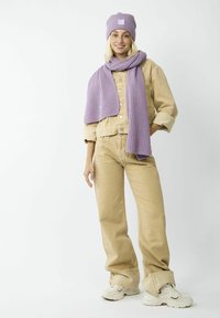 Damen beigefarbene Knopfjacke über beigen Hosen, kombiniert mit einem lila gerippten Strickschal und einer Mütze sowie weißen Sneakers.