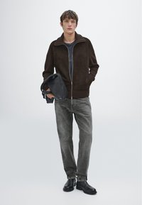 Brązowa kurtka zapinana na zamek z ryflowaną teksturą, noszona na ciemnej koszuli, w połączeniu z jasnoszarymi dżinsami i czarnymi botkami za kostkę. Trzyma czarną torbę.