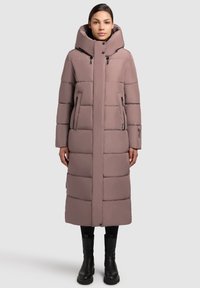 Abrigo largo acolchado rosa con capucha, diseño de bodegas verticales, bolsillos con cremallera y cuello alto. Usado con botas y pantalones negros.