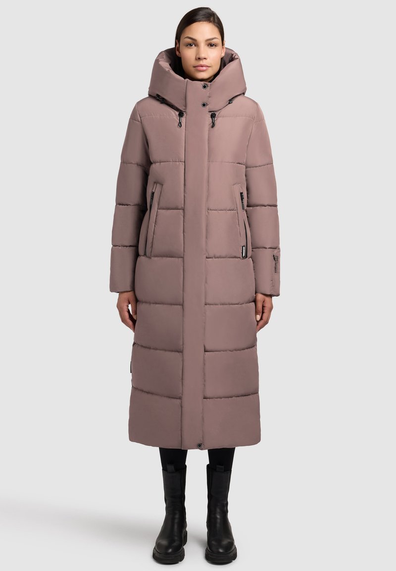 Abrigo largo acolchado rosa con capucha, diseño de bodegas verticales, bolsillos con cremallera y cuello alto. Usado con botas y pantalones negros.