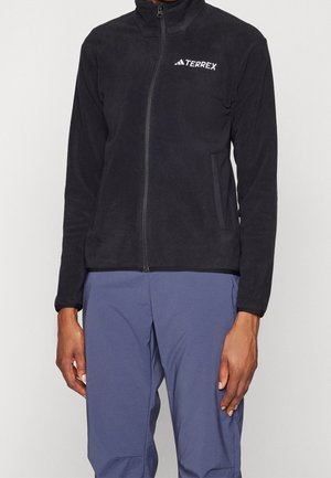 Persoon die een zwarte fleecejas met ritssluiting draagt met Terrex-logo op de borst en blauwe outdoorbroek, staand tegen een effen achtergrond.