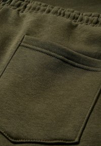 Nahaufnahme von olivgrünem Stoff mit glatter Textur, elastischem Bund und einer einzelnen Patentasche mit verstärkten Nähten.