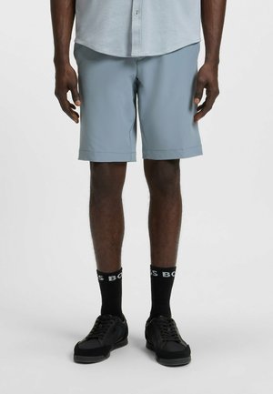 Hellgraue Shorts mit einer glatten Textur, knielang, mit gefalteten Säumen. Getragen mit schwarzen Sneakers und Socken, die das "BOSS"-Logo zeigen.