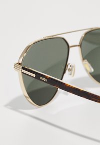 Ochelari de soare aviator aurii cu lentile verzi închise. Brațele templelor din model țestoasă au detalii cu dungi aurii, având imprimat numele brandului „BOSS”.