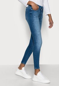 Dunne blauwe denim jeans met een distressed patroon, voorzien van een middelhoge taille en zijzakken, gecombineerd met witte sneakers.