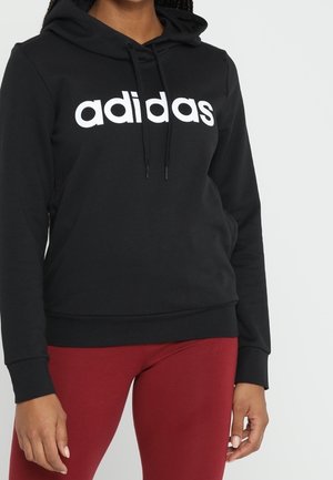 Schwarzer Hoodie mit weißem "adidas"-Logo, Kordelzugkapuze, Fronttasche und gerippten Bündchen. Dazu rote Leggings mit glatter Textur.