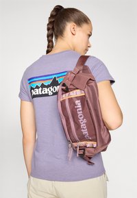T-shirt avec logo Patagonia en violet clair, associé à un sac à dos en bandoulière bordeaux avec des fermetures éclair et des sangles d'accent en marron clair.
