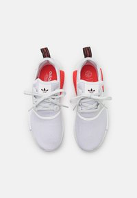 Baltos sportinės avalynės su tinklelio viršumi, raudonais akcentais ir juodu kilpos užtrauktuku. Yra baltos raišteliai ir tekstūruotas padas, užtikrinantis sukibimą.