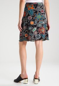 Jupe noire avec des motifs floraux colorés portée par une personne debout, associée à des mules noires à talons hauts en bloc doré.