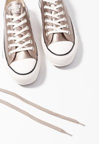 Paire de baskets Converse basses en bronze métallique avec des lacets blancs et des bouts en caoutchouc, à côté d’un lacet beige détaché sur fond blanc.