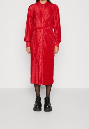 Frau trägt ein rotes Midikleid mit langen Ärmeln, Knöpfen und Taillengürtel, schwarze transparente Strumpfhosen und klobige schwarze Schnürschuhe, steht vor einem einfarbigen Hintergrund.