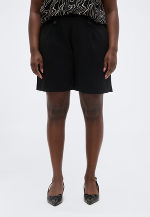 Kobieta w czarnych, wysokich szortach z guzikami, czarnej, wzorzystej bluzce i czarnych butach na obcasie z ostrym noskiem, stojąca na jednolitym tle.