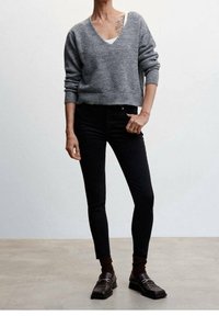 Suéter de lana gris con escote en V, pantalones ajustados negros, calcetines cortos marrones y mocasines marrones oscuros. Presenta un tatuaje visible en el brazo.