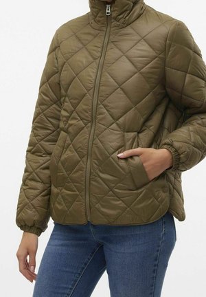 Femme portant une veste matelassée vert olive avec fermeture éclair à l'avant et poignets élastiques, une main dans la poche, associée à un jean bleu.