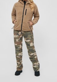 Giacca in pile tan con zip nera e tasche, abbinata a pantaloni cargo mimetici verdi e marroni con tasche laterali e design a gamba dritta.