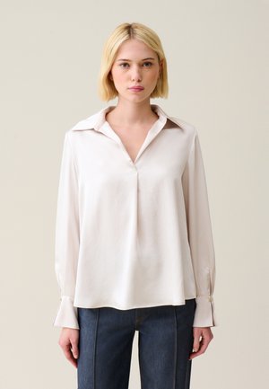Blouse en soie rose clair, avec col, décolleté en V et poignets boutonnés ; présente une coupe ample et une texture lisse. Associée à un jean en denim foncé.
