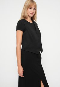 Must have disainiga must vest, millel on V-kaelus, viis nuppu ning lühike pikkus, koos sobiva pika musta seeliku abil.
