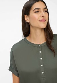 Olijfgroene blouse met een ronde halslijn, korte opgerolde mouwen en een knoopsluiting aan de voorkant met witte knopen. Gladde, lichte stof.