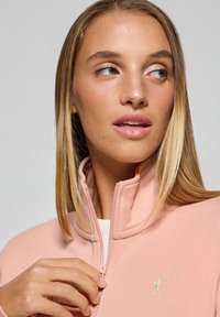 Ljust rosa zip-up jacka med hög krage, tillverkad av mjukt tyg. Har en liten vit logotyp på vänster bröstområde.