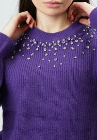 Lila gerippter Pullover mit Rundhals, verziert mit klaren und lila Strasssteinen in verschiedenen Größen im Schulterbereich.