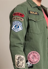 Grön bomullskappa med olika applikationer: "REPLAY," "HERMOSA BEACH SECURITY," och en drake-design. Knappar på framsidan och textilerat tyg.