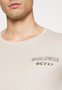 ベージュのコットンTシャツで、クルーネックのデザイン。左胸には黒色の印刷されたテキストデザインで「WORLDWIDEのビジョン」と書かれています。