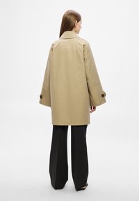 Bézs trench coat nagy, széles ujjakkal, hátul sliccelt, barna gombokkal, fekete bőszárú nadrággal fehér háttér előtt.