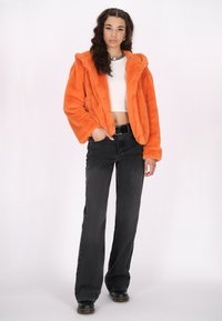 Giacca in pelliccia sintetica arancione con cappuccio, chiusura con bottoni e tasche laterali. Abbinata a jeans neri a vita alta e stivali neri.
