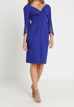 Mujer de pie, vestida con un vestido azul real hasta la rodilla con escote en V y mangas de encaje 3/4, combinado con sandalias plateadas de tacón alto.
