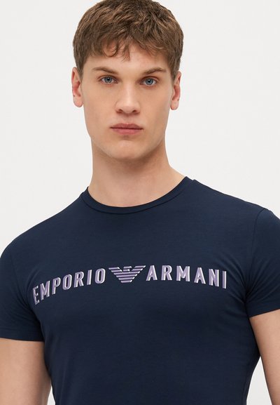 T-shirt ajustado azul-marinho feito de algodão macio, com o logótipo 'EMPORIO ARMANI' em tons de roxo degradê na parte da frente. Gola redonda.