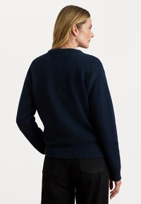 Marineblauwe gebreide trui met een ronde hals, geribbelde zoom en manchetten. De stof heeft een getextureerde afwerking. Draagt over zwarte broek.