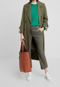 Olivově zelený dvouřadový trenčkot, tyrkysový svetr, šedé kalhoty na míru, oranžová texturovaná kabelka a bílé tenisky.