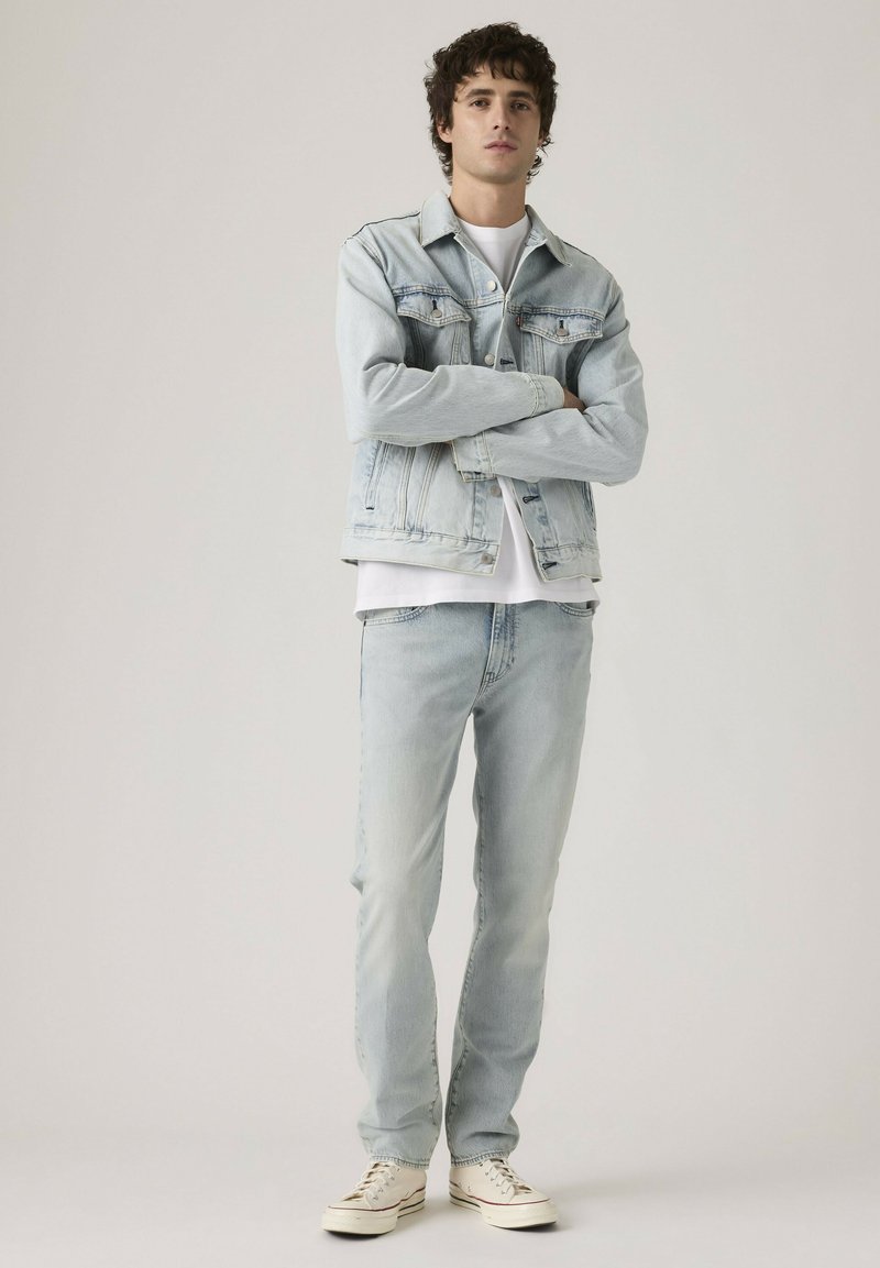 Young man standing with arms crossed wearing light blue denim jacket, matching jeans, white t-shirt, and off-white sneakers.