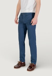 Chinos blu in cotone da uomo con una vestibilità slim, dotati di tasche frontali, chiusura a bottone e una texture liscia, abbinati a scarpe marroni.