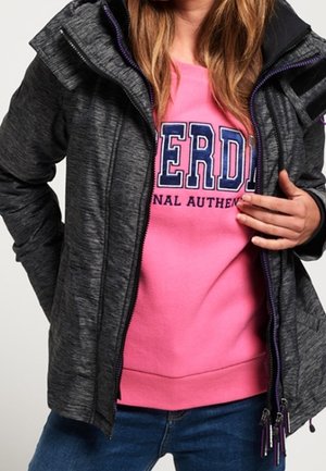 Grå zip-up jakke med en struktureret finish og lilla detaljer, lagret over en lyserød sweatshirt med fed tekst. Jeans fuldender outfittet.