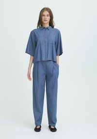 Jonge vrouw staat, gekleed in een losse blauwe denim knopenoverhemd en bijpassende hoge taille broek met zwarte platte schoenen, tegen een eenvoudige achtergrond.