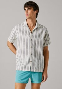 Ung mand iført en hvid kortærmet skjorte med grå lodrette striber og turkis shorts, stående mod en ensfarvet baggrund.