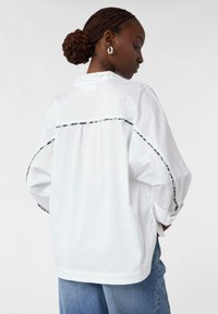 White long-sleeve shirt with black trim along the back and sleeves. Features a relaxed fit and round hem. Made of smooth fabric.