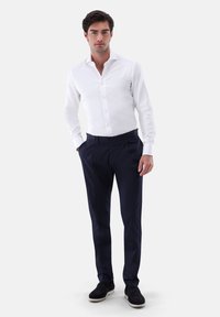 Camicia bianca con un motivo testurizzato, abbinata a pantaloni blu navale con pince frontali. Il modello indossa sneaker scure.