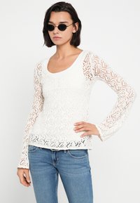 Λευκό πλεκτό τοπ με μακριά μανίκια και υφασμένο σχέδιο δαντέλας, στρογγυλή λαιμόκοψη και μανσέτες μανικιών που ανοίγουν προς τα έξω, φορεμένο με μπλε τζιν παντελόνια.