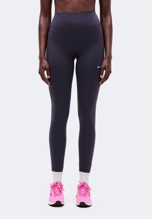Modèle portant un legging foncé taille haute, des chaussettes blanches et des chaussures de course rose vif, sur un fond uni.
