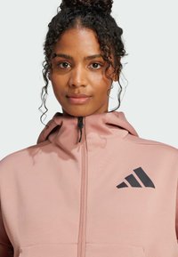 Sudadera con cremallera de color rosa claro, con capucha, tela texturada y un logotipo negro de Adidas en el lado izquierdo del pecho. Se puede ver el detalle del cierre de cremallera.