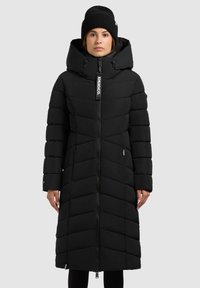 Crna puf jakna s prednjim zatvaračem, visokim ovratnikom i kapuljačom. Ima šavove u obliku češljane crte i bočne džepove. Uključuje oznaku s logotipom na vratu.