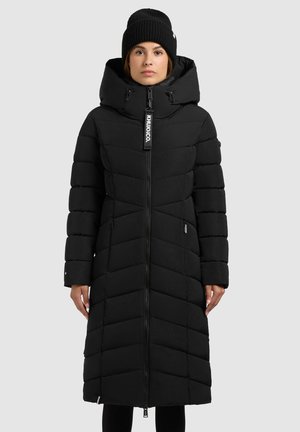 Juodas pūkinis paltas su užtrauktuku priekyje, aukštu apykakle ir gobtuvu. Yra čečėnų siuvimo elementai ir šoninės kišenės. Kaklo srityje yra logotipo etiketė.