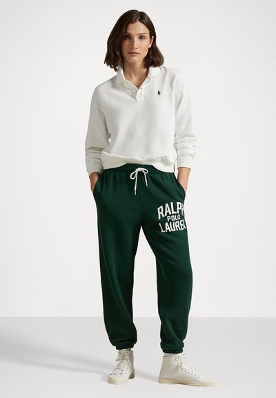 Biely polo s dlhým rukávom a golierom v kombinácii s tmavozelenými teplákmi so sviežim bielym textom "RALPH LAUREN" a svetlou obuvou.