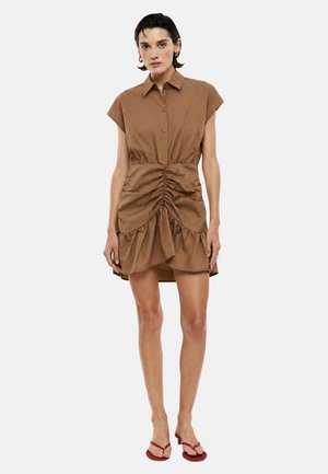 Donna con capelli corti e scuri che indossa un vestito marrone con maniche corte arricciate e sandali rossi con punta aperta, in piedi su uno sfondo semplice.