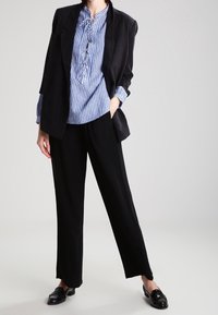 Blazer noir ajusté par-dessus une blouse rayée bleue et blanche avec un devant à lacets, associé à un pantalon large noir et des mocassins noirs.
